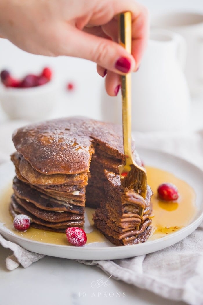 Stack of paleo gingerbread pancakes on a white plate with maple syrup around the bottom and a big bite cut out with a gold fork held between fingers in the piece and fresh cranberries as garnish