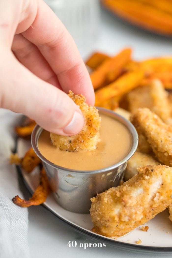 Hand dipping paleo Whole30 chicken nuggets into dipping sauce sauce with sweet potato fries in background