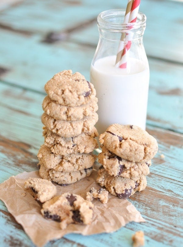 Paleo Chocolate Chip Cookies stacked on top of each other on brown parchment paper leaning against a glass of milk with two straws sticking out.