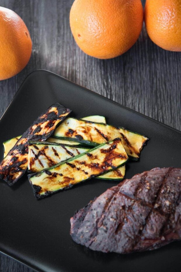 Overhead shot of grill-marked orange glazed grilled zucchini slices arranged on a black plate next to a steak with some oranges in the background.