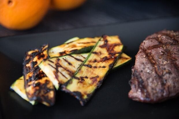 Close-up of grill-marked orange glazed zucchini slices arranged on a black plate next to a steak with oranges in the background.