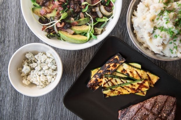 Overhead shot of grill-marked orange glazed grilled zucchini slices arranged on a black plate next to a steak with more BBQ side dishes in the background.