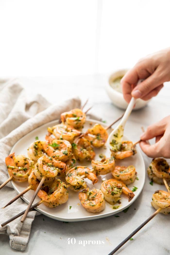Hands drizzling chimichurri sauce on chimichurri shrimp skewers, piled on a plate