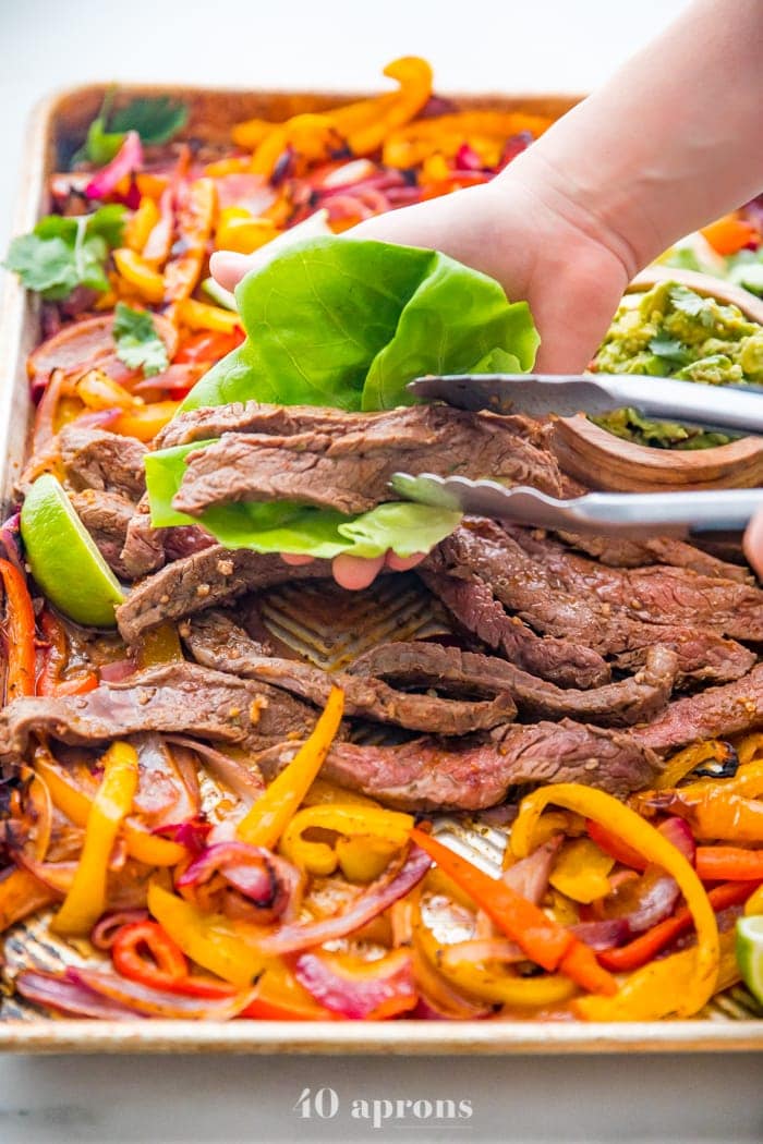 Best sheet pan fajitas with steak and peppers on a sheet pan with a hand putting steak in a lettuce cup