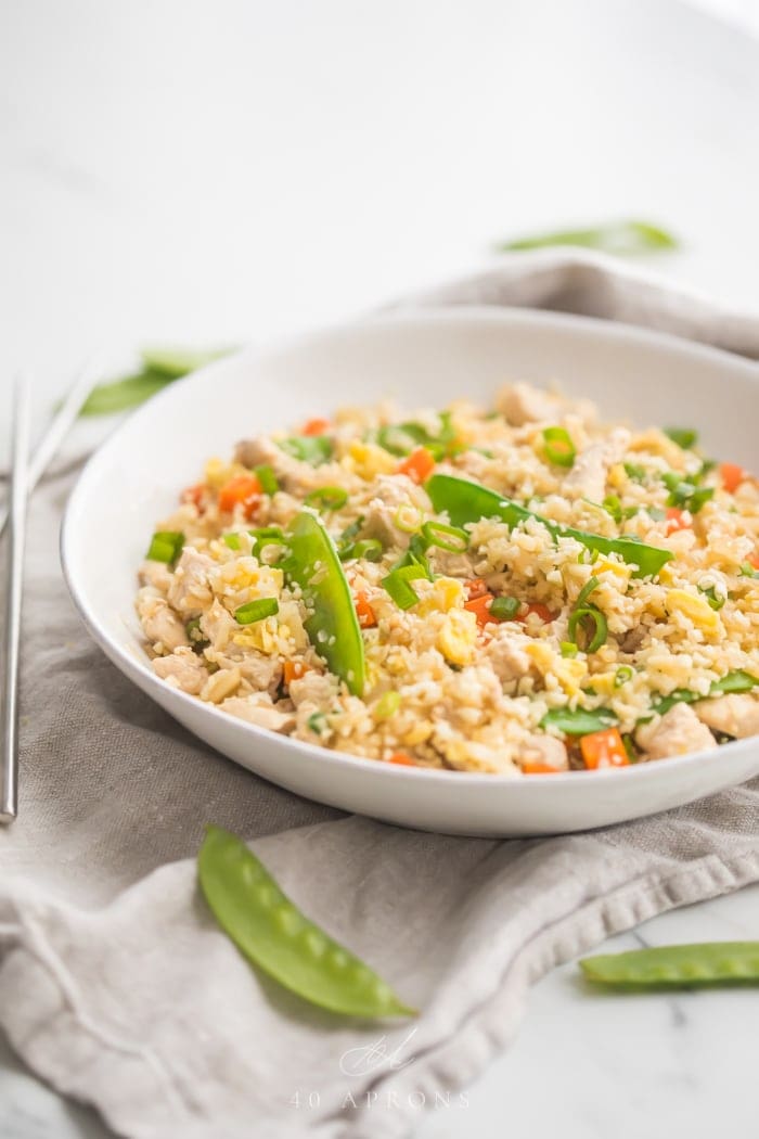 A white bowl of cauliflower fried rice with chicken, peas, carrots with a grey linen and silver chopsticks to the side