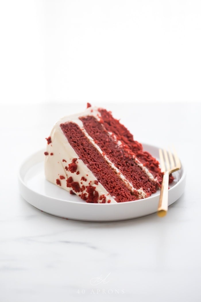 Slice of three layer gluten free red velvet cake with paleo cream cheese frosting on a white plate with a gold fork