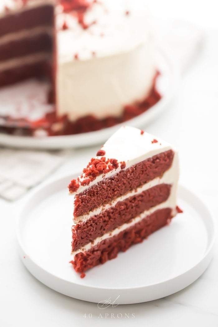 Slice of gluten free red velvet layer cake with paleo cream cheese frosting on a white plate with whole cake in background