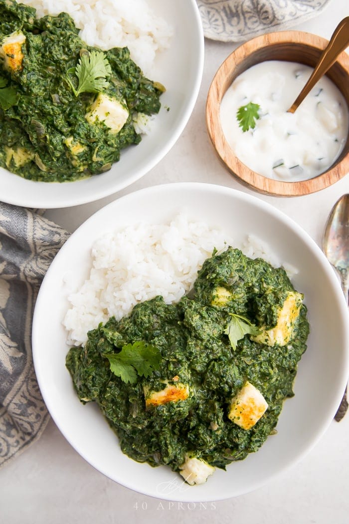 Two bowls of palak paneer over rice with raita on the side