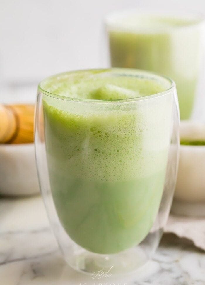 two matcha lattes in glass cups with a bamboo whisk to the side