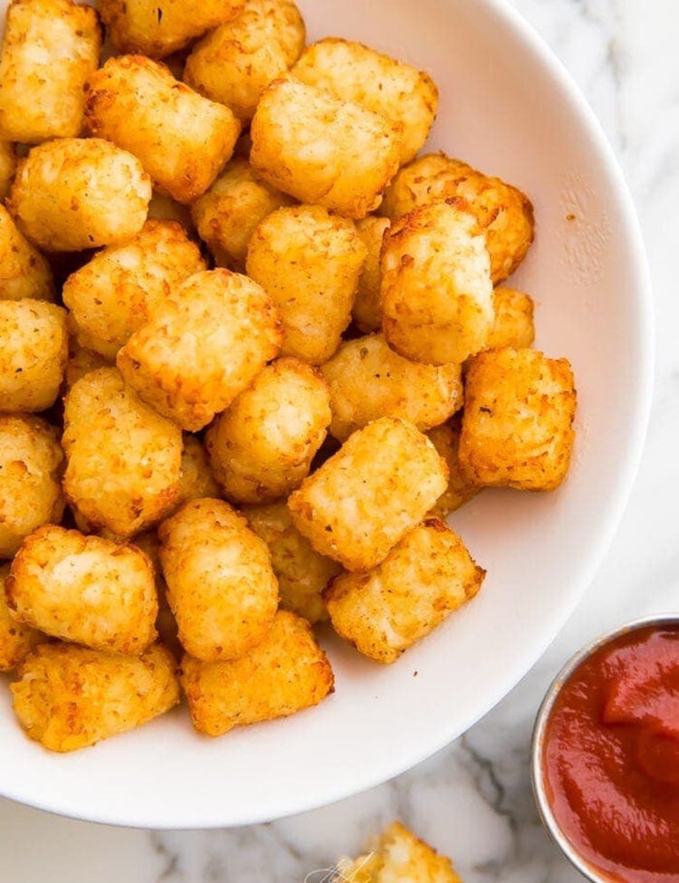 Crispy, golden air fryer tater tots in a large white serving bowl next to a small ramekin of bright red ketchup.