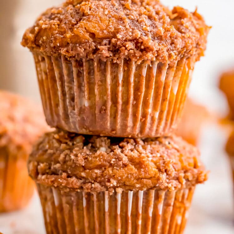 Two pumpkin muffins with a spiced crumb topping stacked on top of each other, with other muffins out of focus in the background.