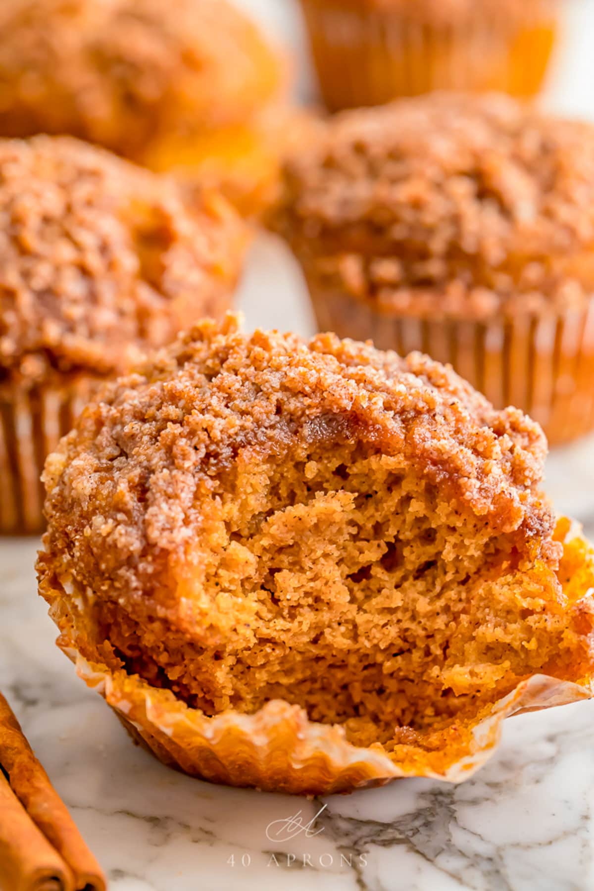 A pumpkin muffin, with the paper lining peeled down and a large bite missing from the front side of the muffin. More whole muffins stand in the background.