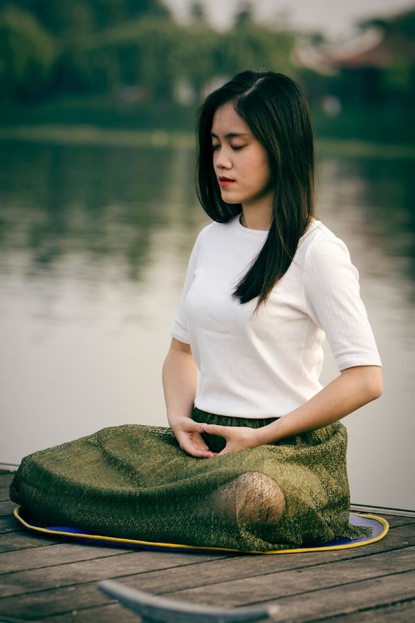 a young Asian woman meditating sitting in the lotus position on the pier of a lake 