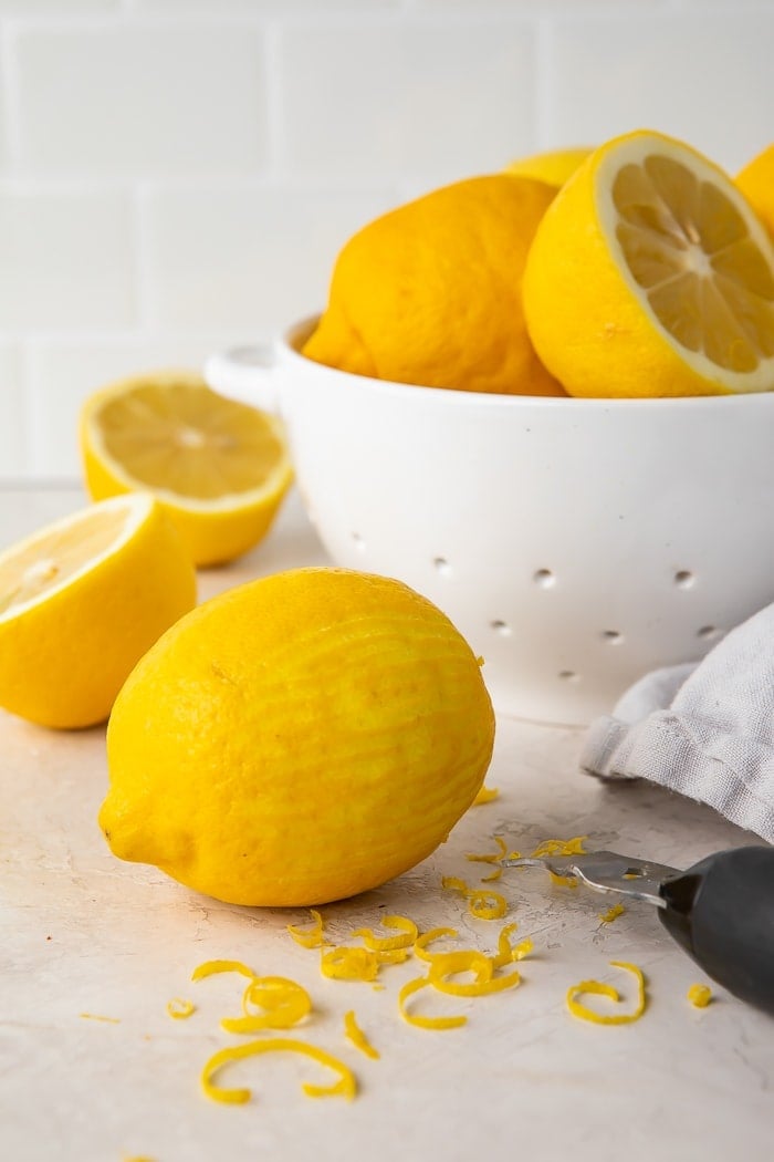 a zested lemon surrounded by lemon zest, a zester, and a colander of lemons