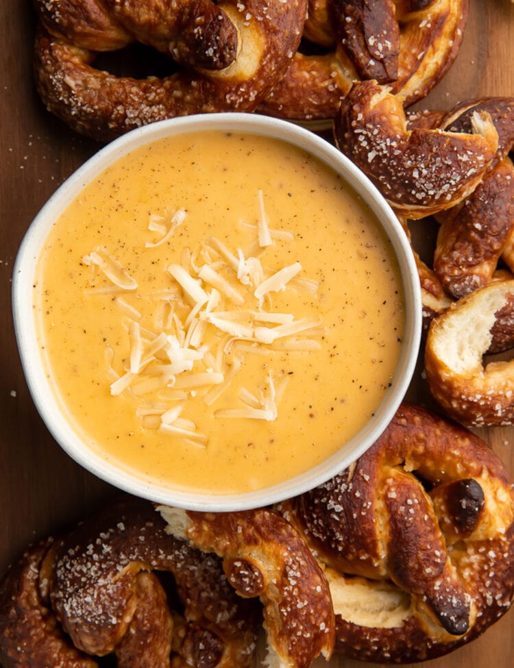 Overhead, top down look at a white bowl of rich yellow-orange beer cheese dip topped with shredded mozzarella surrounded by deep brown soft pretzels with salt.