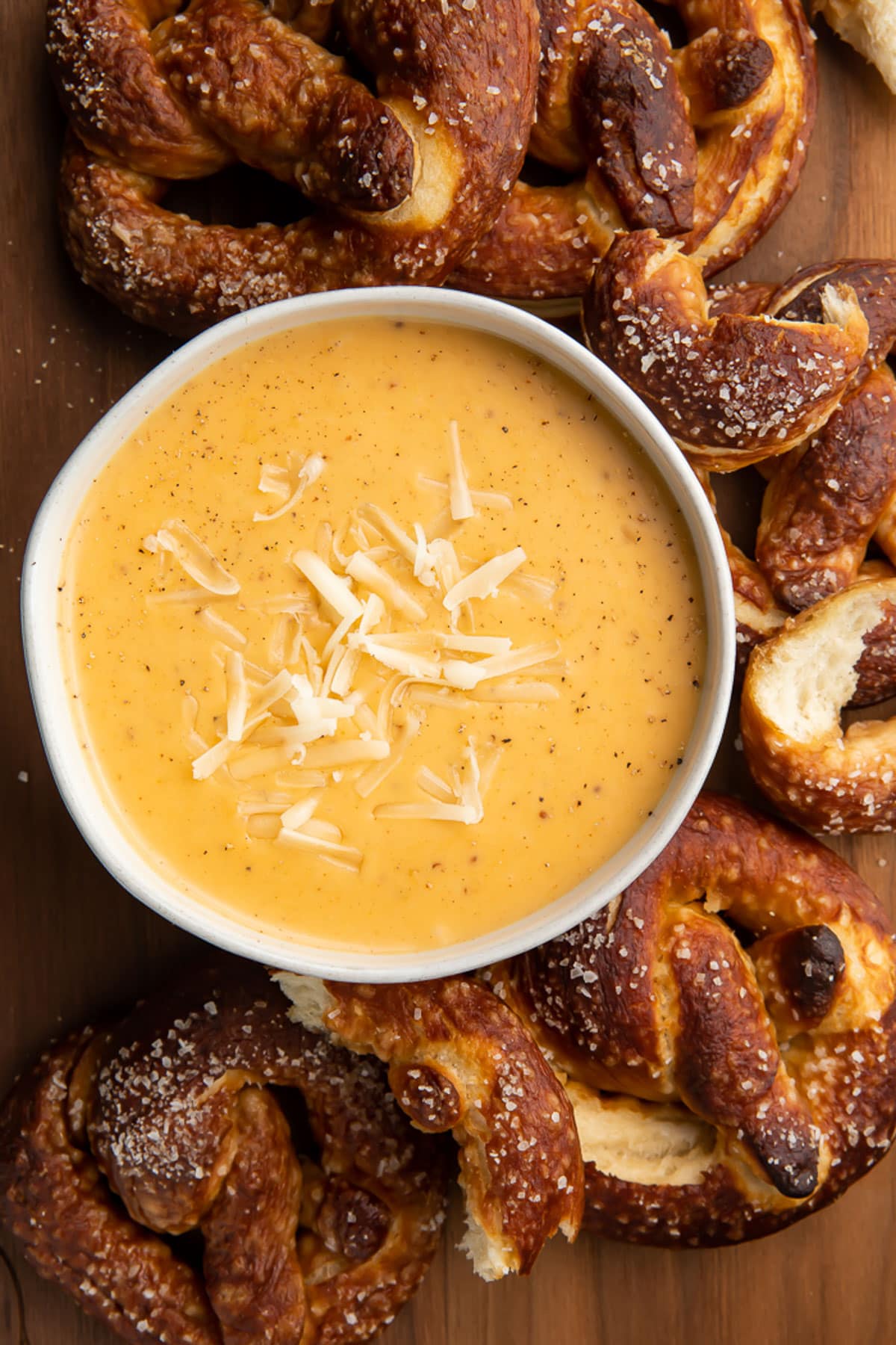 Overhead, top down look at a white bowl of rich yellow-orange beer cheese dip topped with shredded mozzarella surrounded by deep brown soft pretzels with salt.