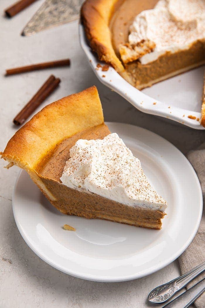 A slice of keto pumpkin pie topped with whipped cream on a white plate with a pie in the background