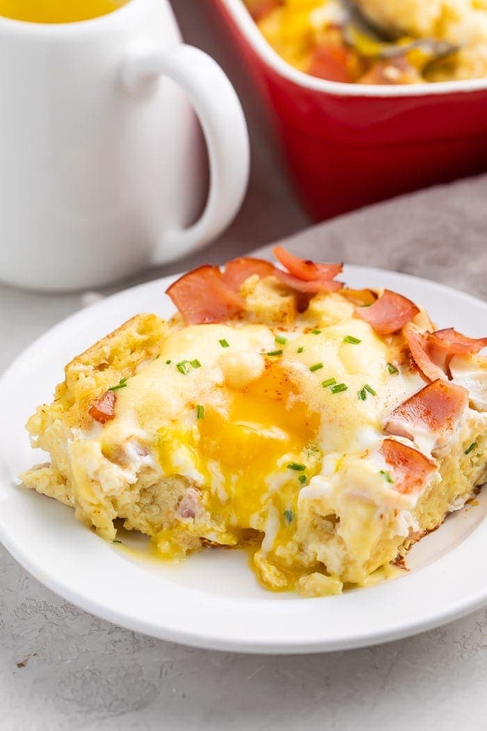 A slice of eggs benedict casserole with a bite taken out on a white plate with a mug and red bowl in the background