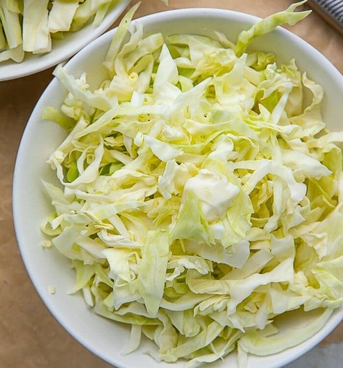 A white bowl full of shredded cabbage on a wooden cutting board next to a knife
