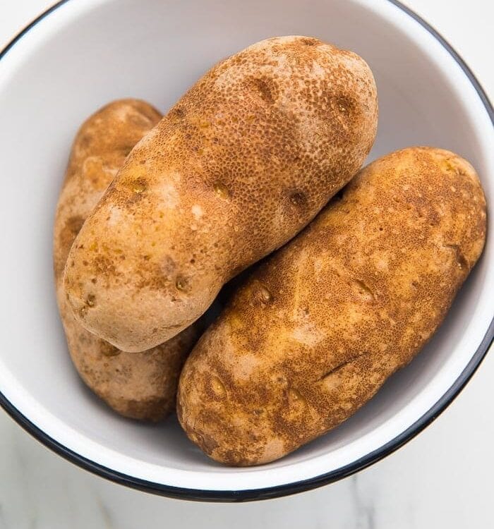 3 unpeeled russet potatoes in a black rimmed white bowl on a marble countertop
