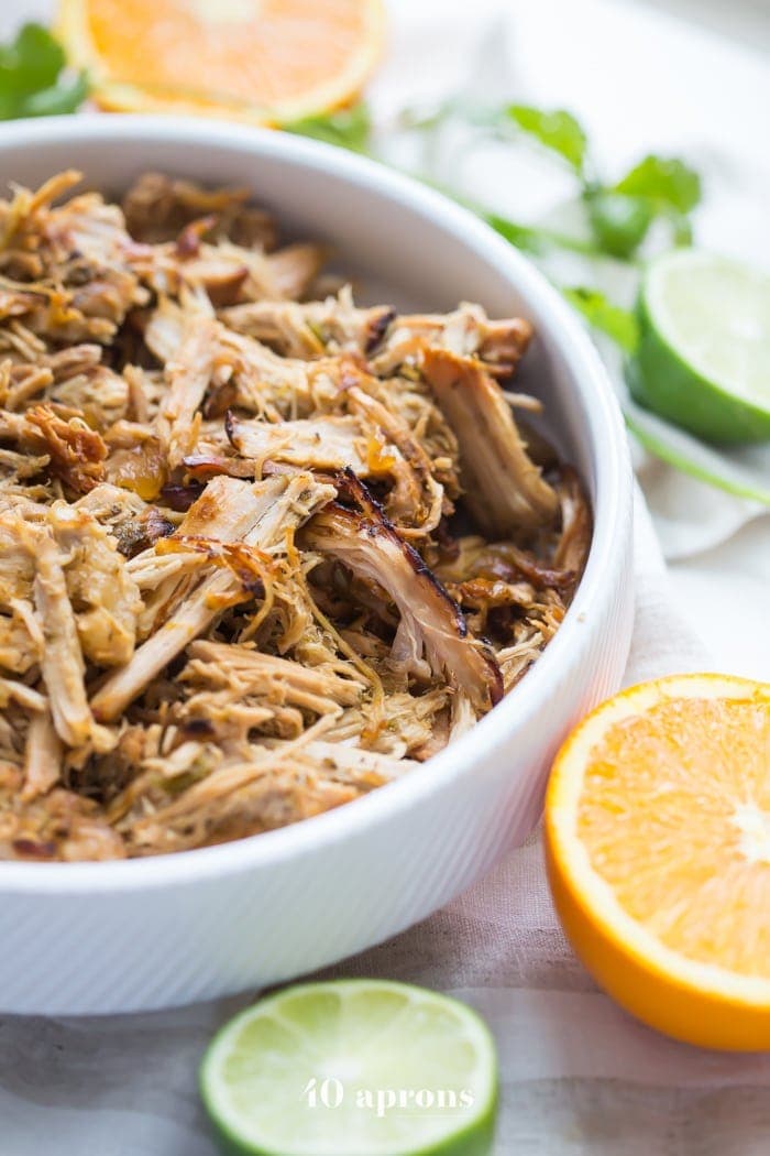 A white bowl with carnitas cooked in a Crockpot surrounded by cut citrus