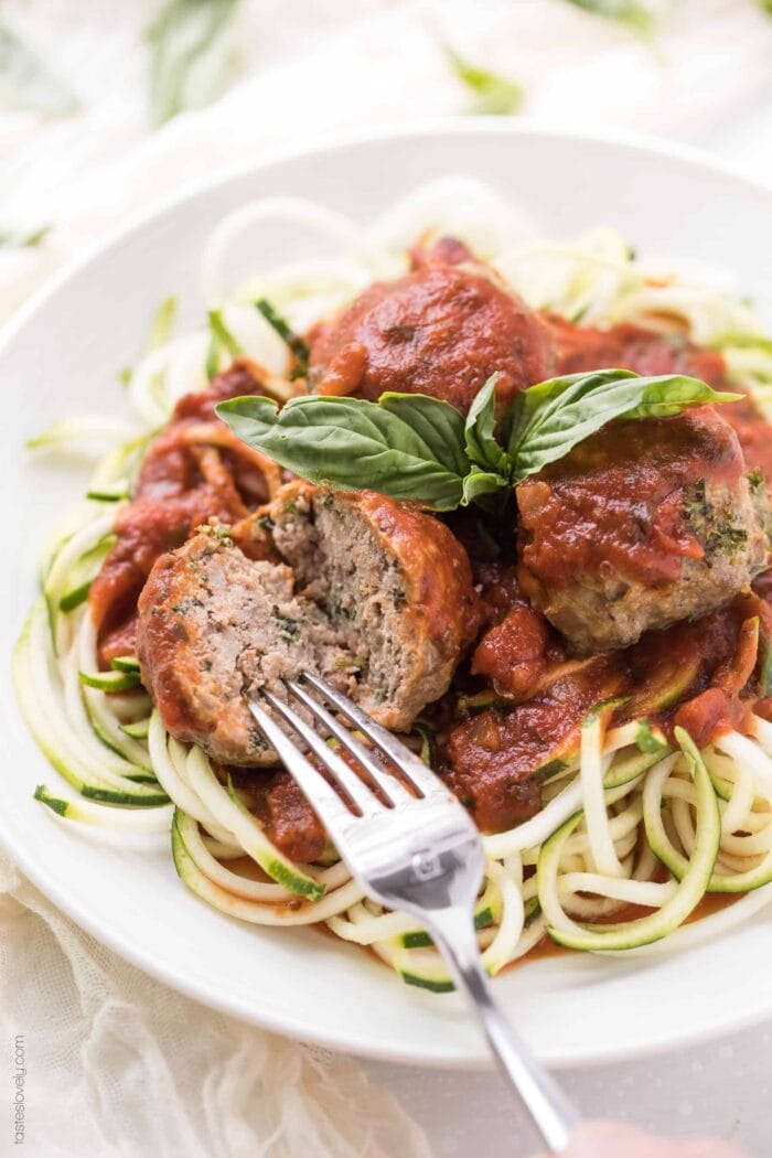 Whole30 Italian meatballs on a bed of zoodles. One meatball is being cut open with a fork.