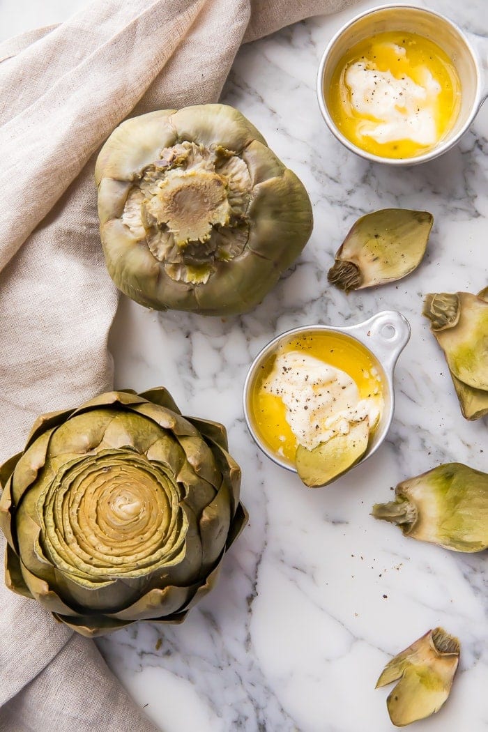 Two artichokes on a marble counter with bowls of dipping sauce
