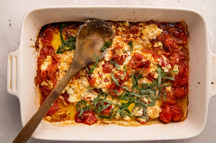 Baked feta and tomatoes, mashed together, with basil and olive oil in a casserole dish, with a wooden spoon laid across diagonally