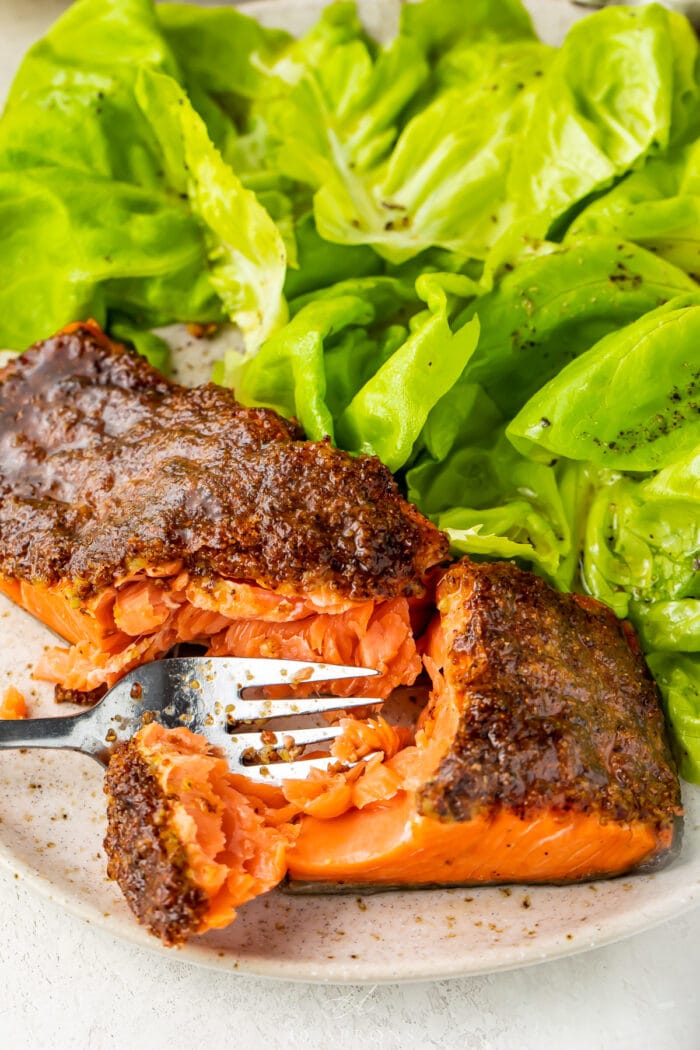 A fork cutting through flaky air fryer salmon on a plate with a small green salad