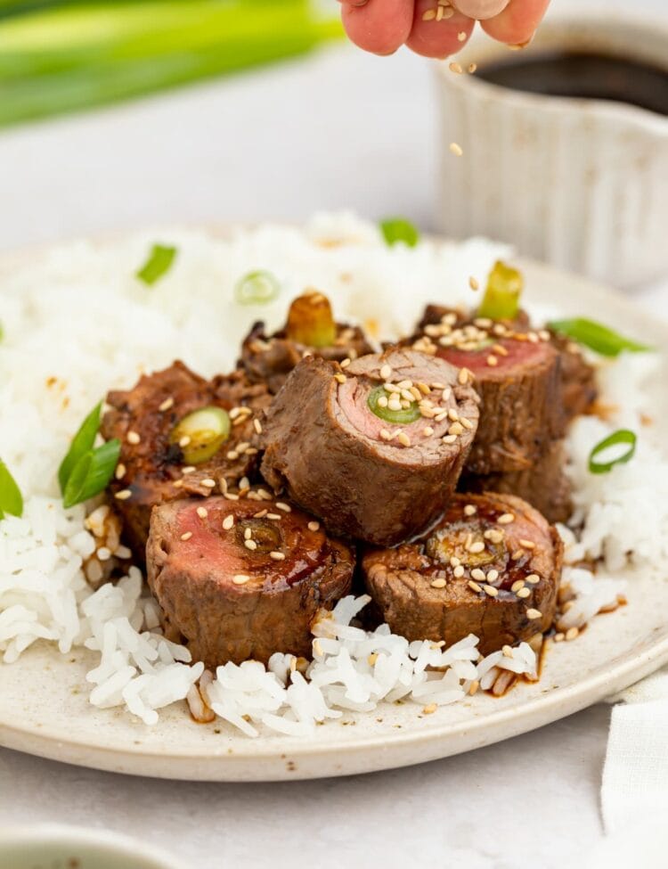 beef negimaki on a plate with rice and sesame seeds being sprinkled over the top