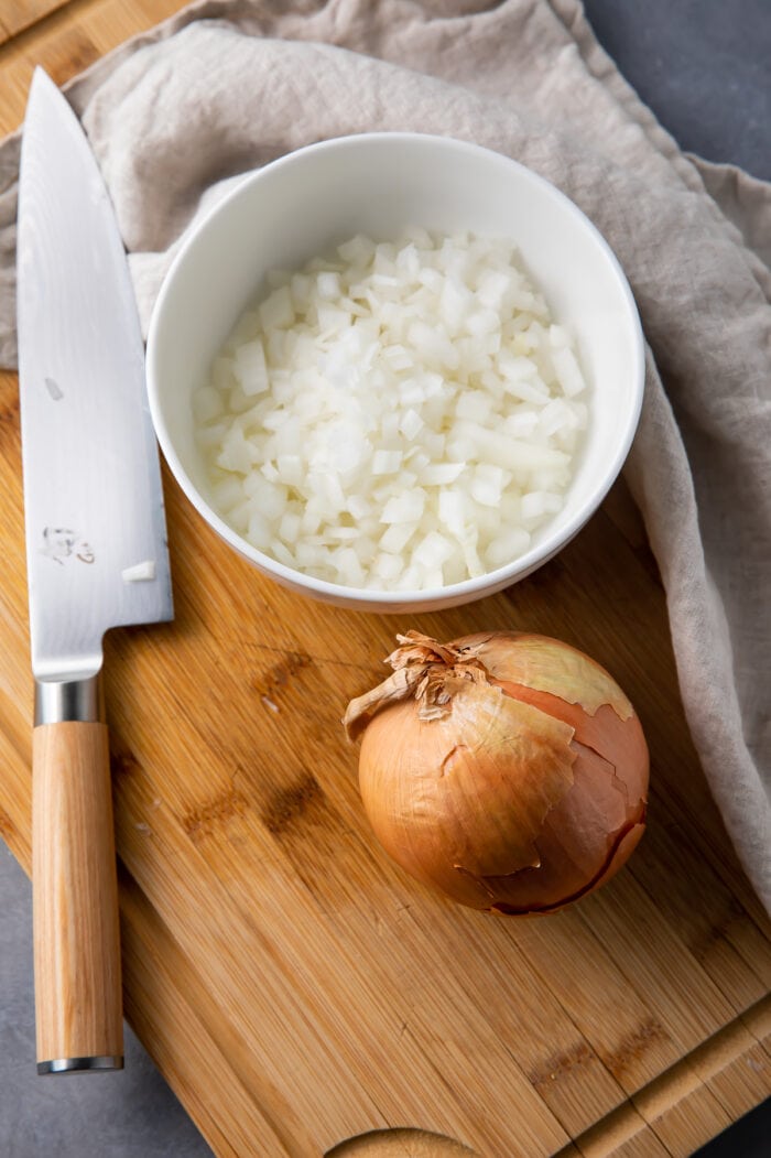A whole onion next to a bowl of diced onion and a knife on a cutting board with a dish towel