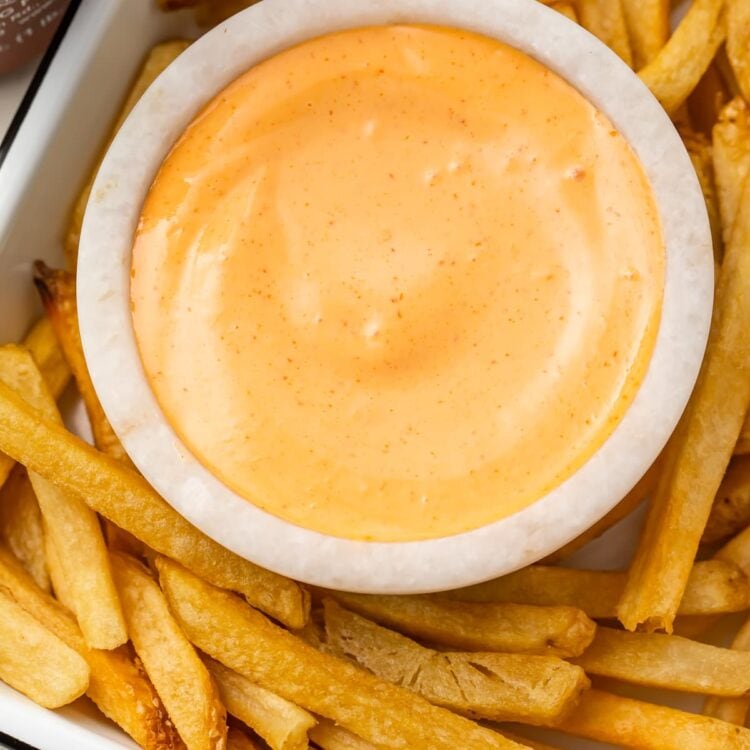 A small bowl of sriracha mayo on a baking sheet surrounded by french fries