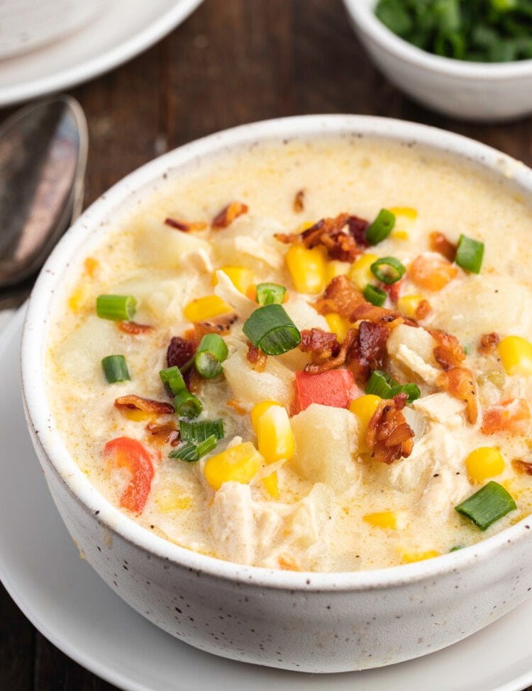 A white bowl of chicken corn chowder sitting on a white plate on top of a dark wooden table.
