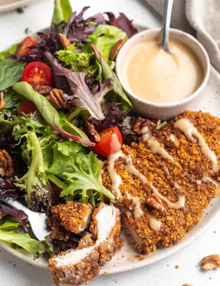 Close-up, angled view of pecan crusted chicken with honey mustard sauce and small side salad on a white plate.