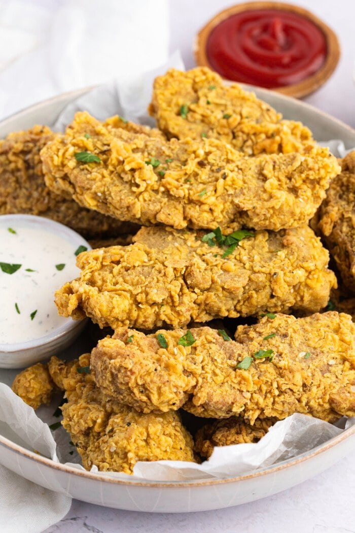 A pile of vegan seitan fried "chicken" tenders in a white bowl next to a small ramekin of ranch dressing with a small ramekin of ketchup in the background.