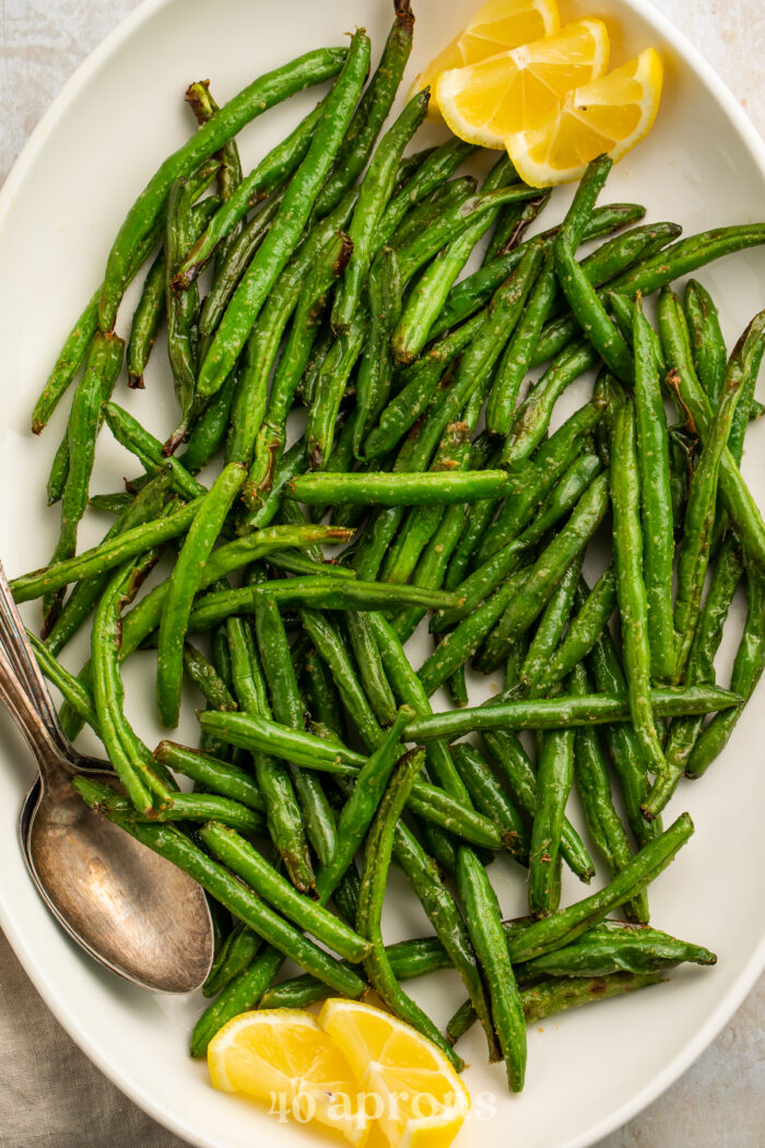 Close-up of air fryer green beans on a white serving platter with lemon wedges and a silver spoon.