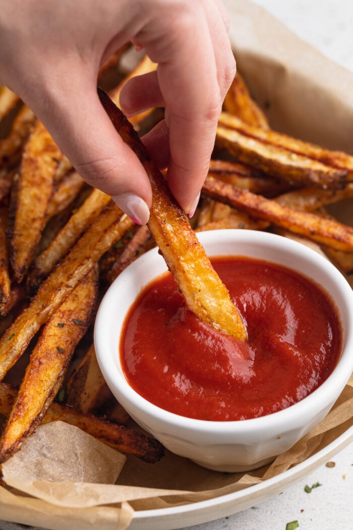 A cajun fry being dipped in a ramekin of ketchup resting on a plate with more cajun fries.