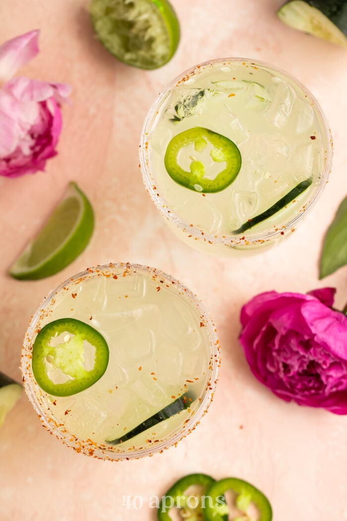Overhead view of two cucumber jalapeno margaritas on a table with cucumber slices and fuschia flowers.