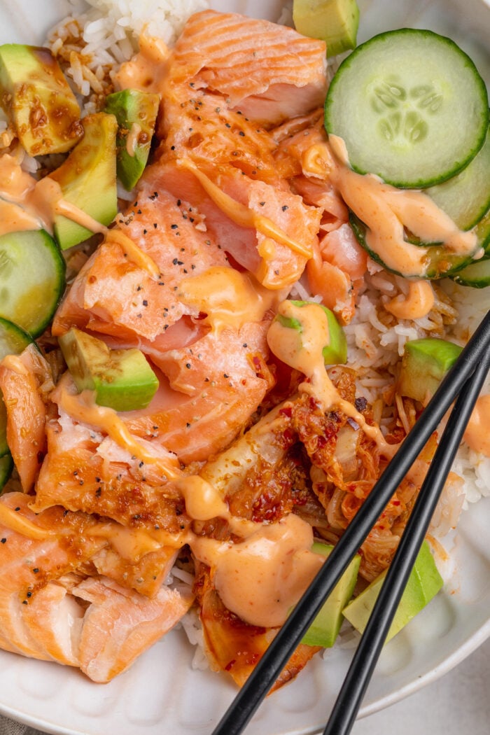 Close-up view of oven-baked salmon fillets with white rice, soy sauce, creamy sriracha mayo, cucumber, avocado, and chopsticks.