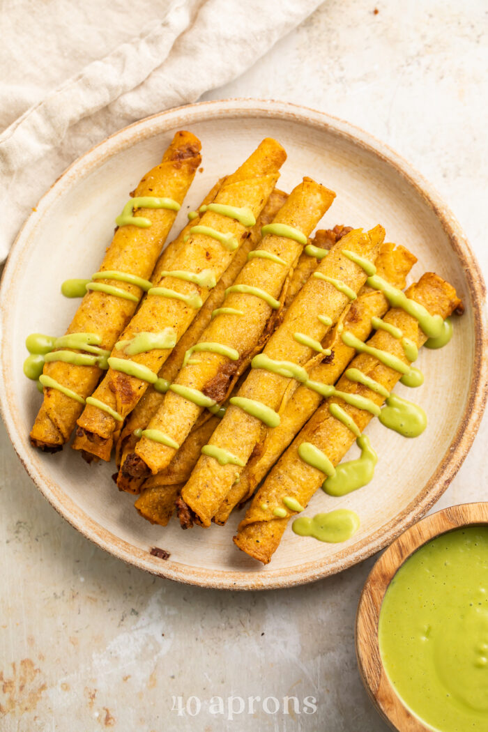 Overhead view of crispy frozen taquitos cooked in the air fryer on a plate, with a drizzle of avocado sauce.