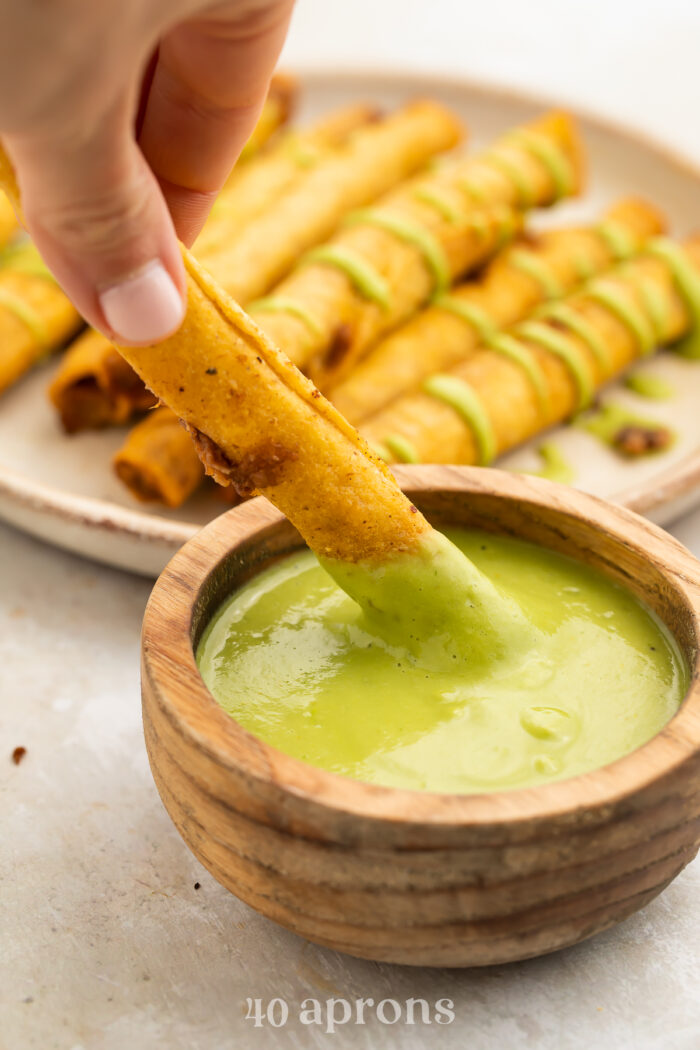 A crispy, air fryer frozen taquito being dipped into a small wooden bowl filled with a creamy avocado sauce.
