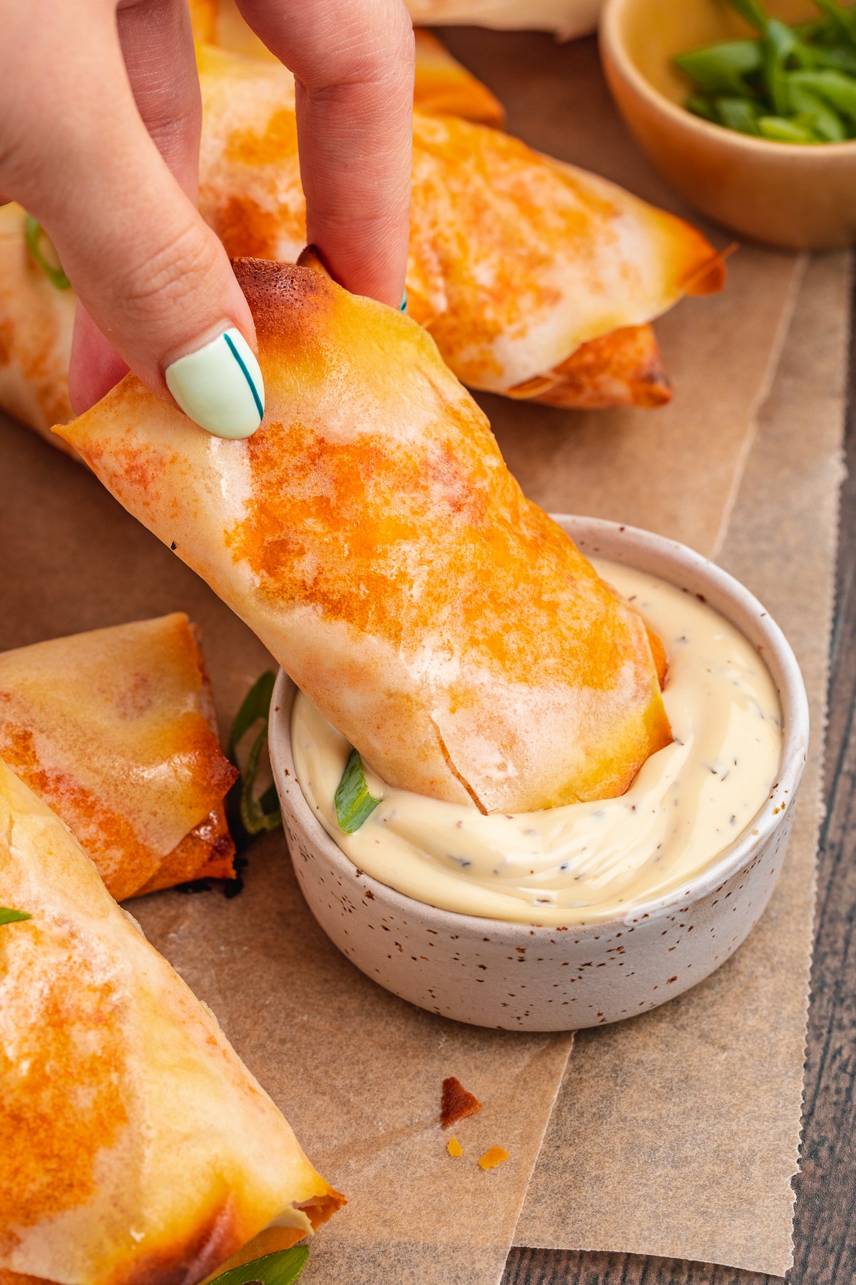 A white woman's hand with painted nails dipping a buffalo chicken egg roll into a small ramekin of ranch dressing on a baking sheet lined with parchment paper.