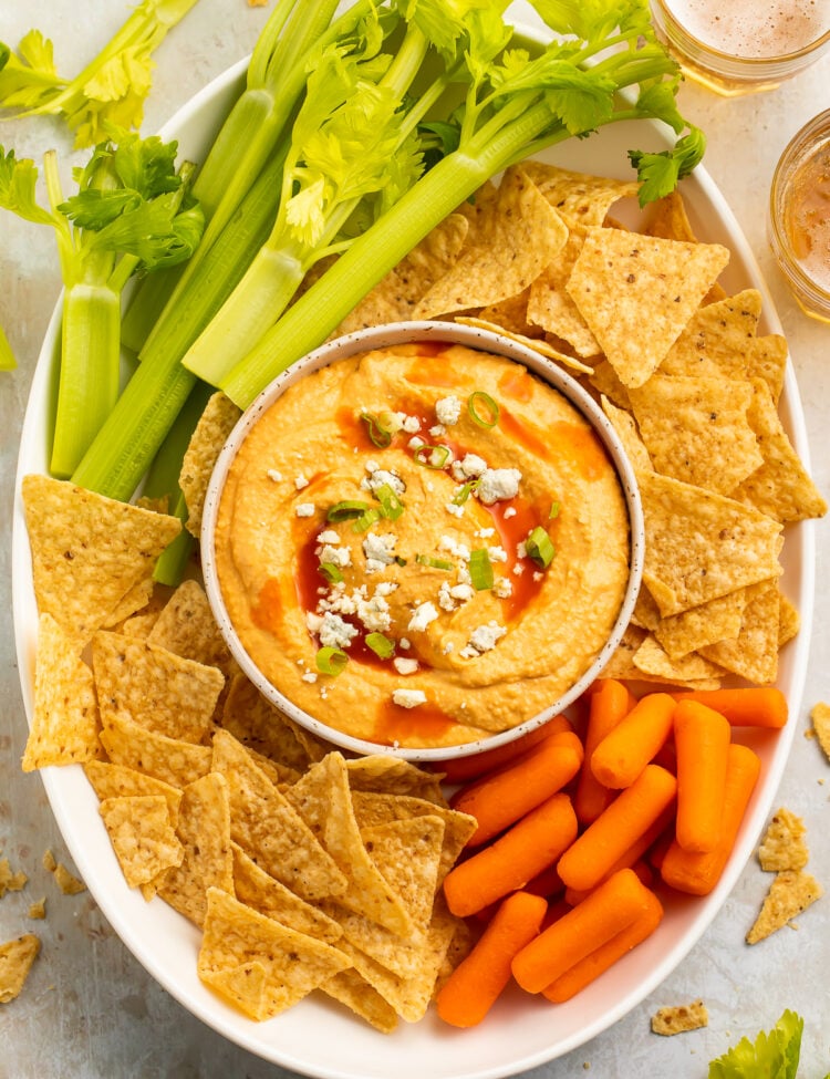 Overhead view of a bowl of buffalo hummus topped with blue cheese crumbles in the center of a platter surrounded by celery, carrots, and tortilla chips.