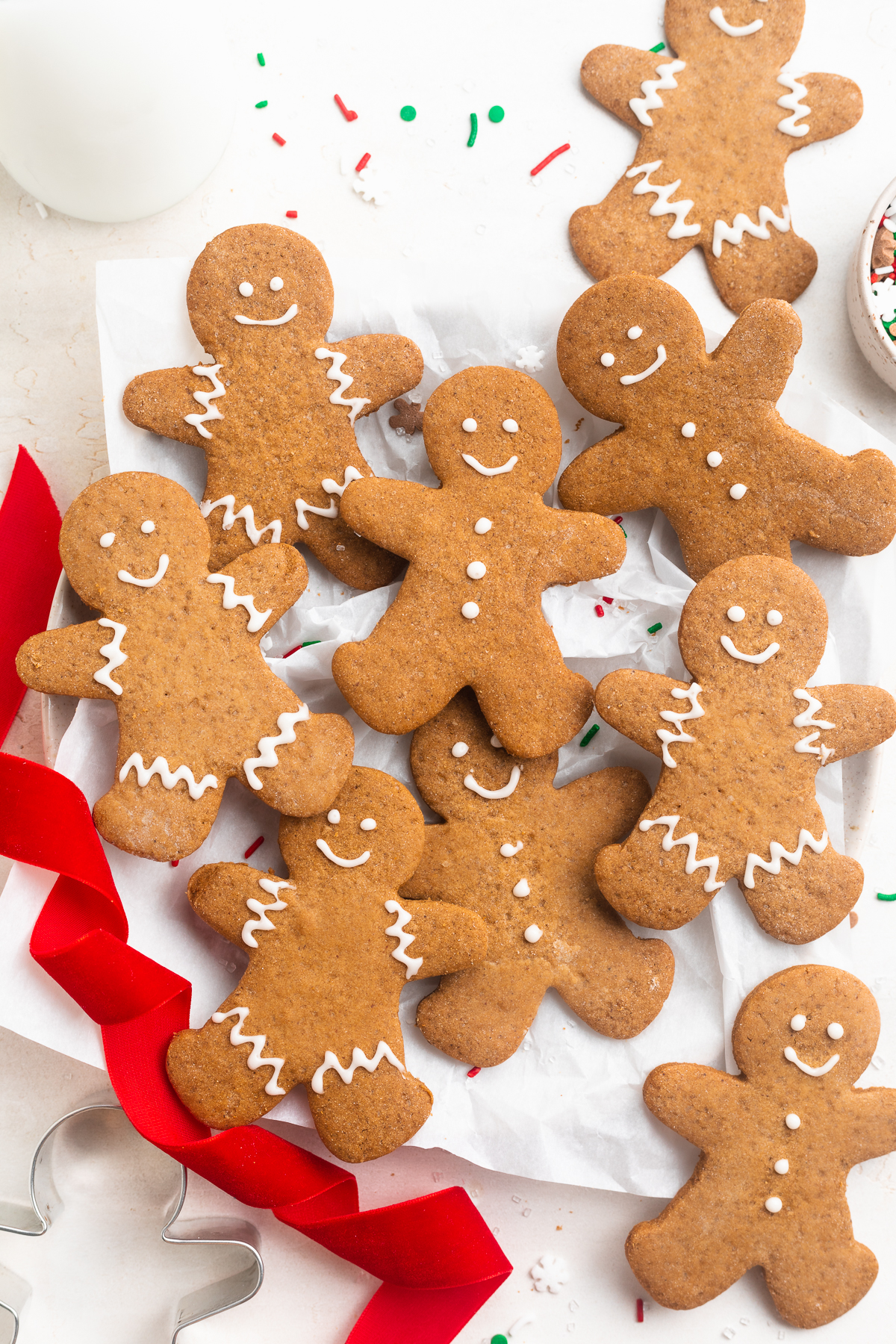 Overhead photo of gluten-free gingerbread men decorated with white cookie icing on a white table with red ribbon and holiday sprinkles.