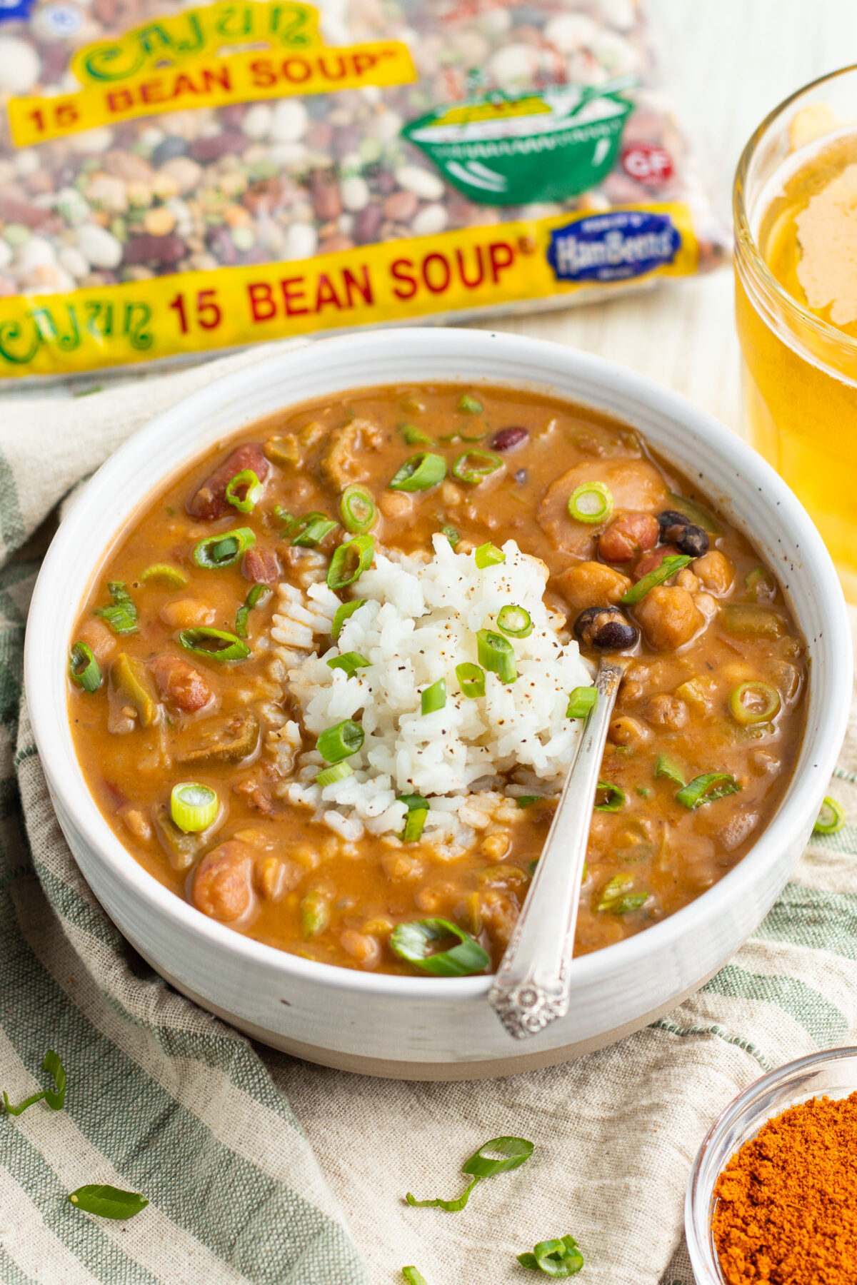 A white bowl filled with 15-bean vegetarian gumbo, topped with white rice and slices of green onions, with a Hurst's HamBeens Cajun 15-Bean Soup mix bag in the background.