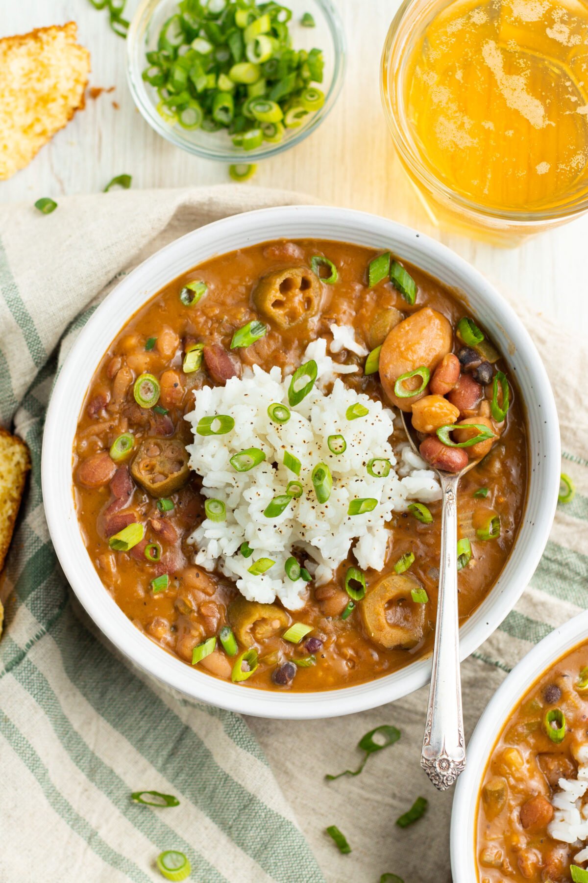 Overhead photo of a white bowl filled with reddish-brown Cajun 15-bean vegetarian gumbo, topped with rice and slices of green onion.