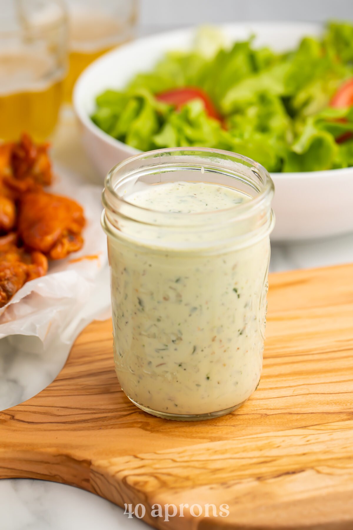 A mason jar of ranch dressing peppered with green specks from fresh herbs sits on a wooden cutting board, in front of a bright green salad and orange chicken wings.