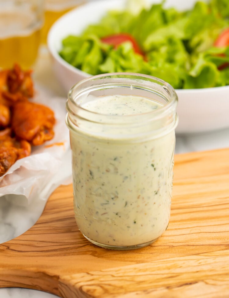 A mason jar of ranch dressing peppered with green specks from fresh herbs sits on a wooden cutting board, in front of a bright green salad and orange chicken wings.
