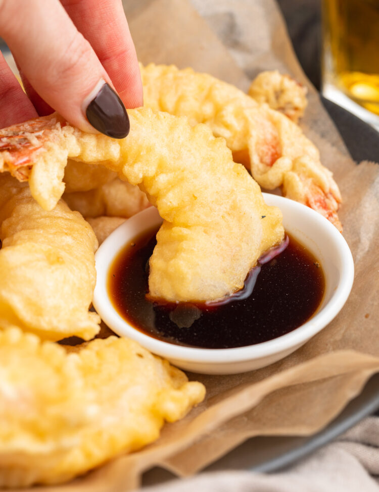 A large piece of shrimp tempura being dipped in a small bowl of soy sauce by a white woman's hand with black painted fingernails.