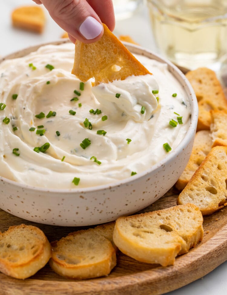 A white woman's hand using a crostini to scoop creamy boursin dip out of a white bowl.